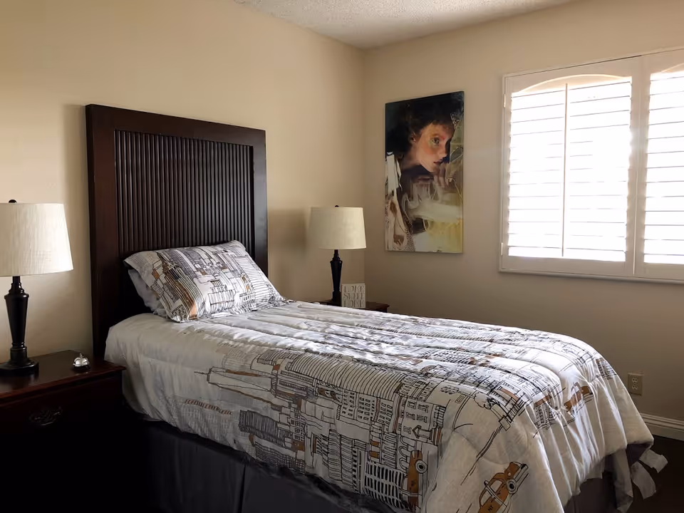 A neatly made single bed with a dark wooden headboard and cityscape-themed bedding in a bedroom. There are two bedside tables, each with a lamp, and a painting of a person's face on the wall next to a window with white shutters.