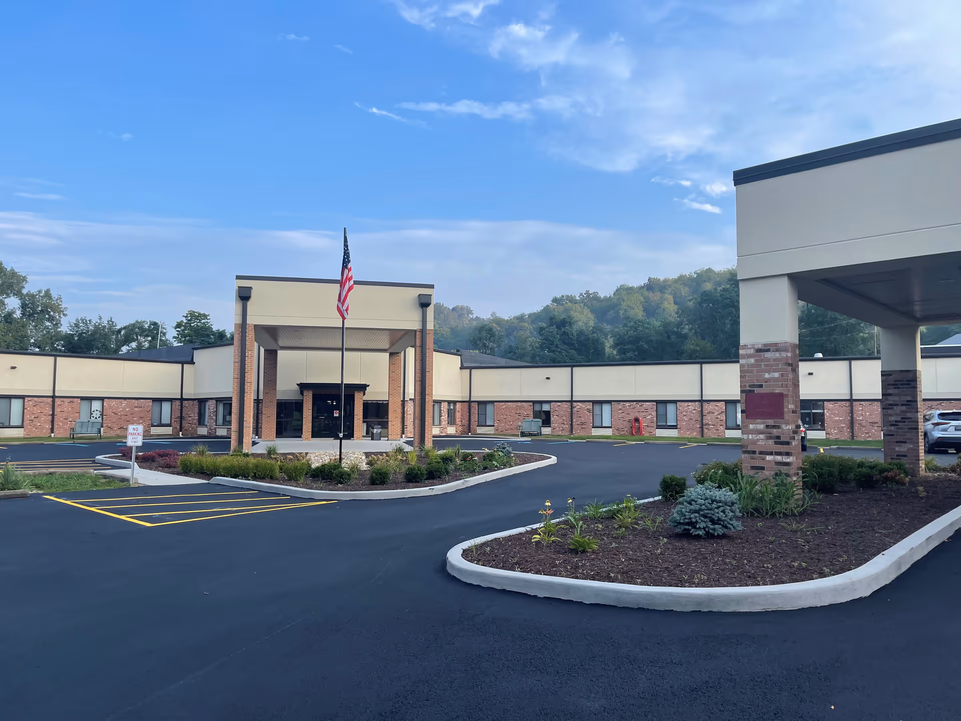 Exterior view of Moundsville Healthcare Center showing a single-story building with a covered entrance, an American flag on a flagpole, landscaped areas with plants, and a paved driveway under a partly cloudy sky.