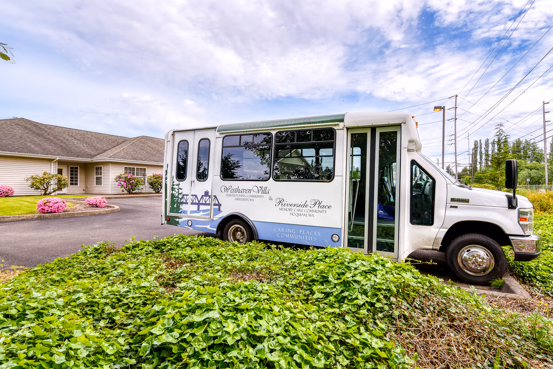 A white shuttle bus parked outside a single-story building with a well-maintained garden featuring green shrubs and pink flowers. The bus has signage for Westhaven Villa Assisted Living Community and Riverside Place Memory Care Community.