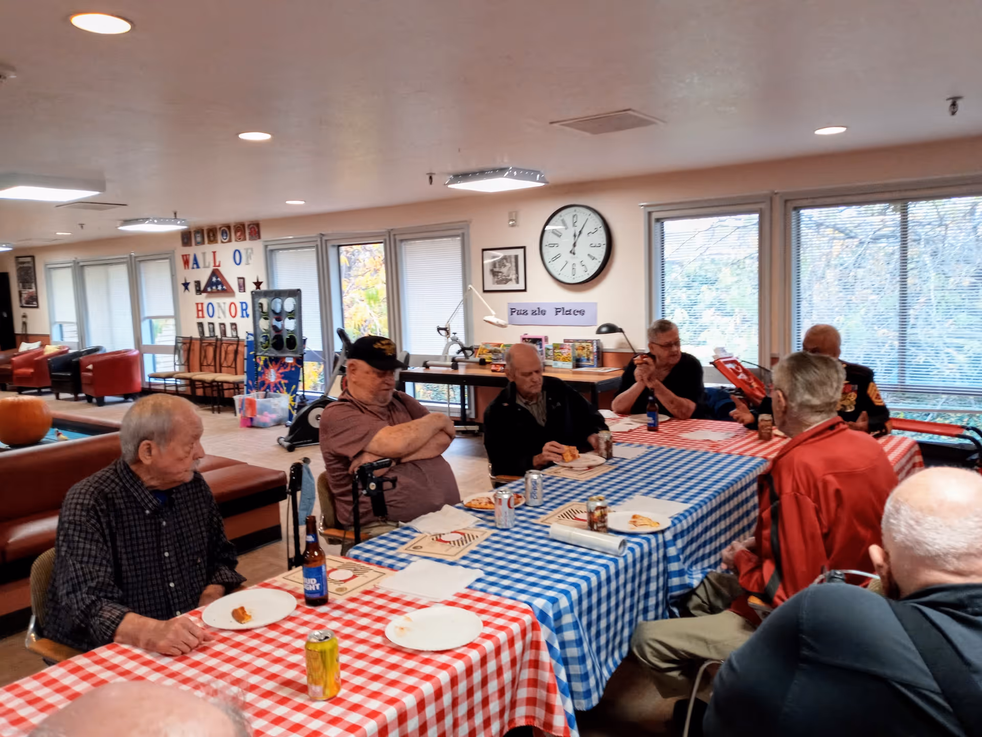 A group of elderly men sitting around two tables covered with red and blue checkered tablecloths, eating pizza and drinking soda in a well-lit room with large windows. The room has a clock on the wall, a sign that reads 'Puzzle Place,' and a 'Wall of Honor' display in the background.
