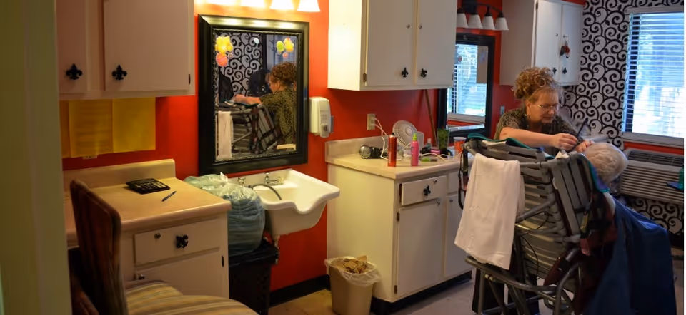 A caregiver styling the hair of an elderly person seated in a wheelchair in a small room with red walls, white cabinets, a sink, and a mirror. The room has a window with blinds and patterned curtains, and various hair care products are placed on the counter.