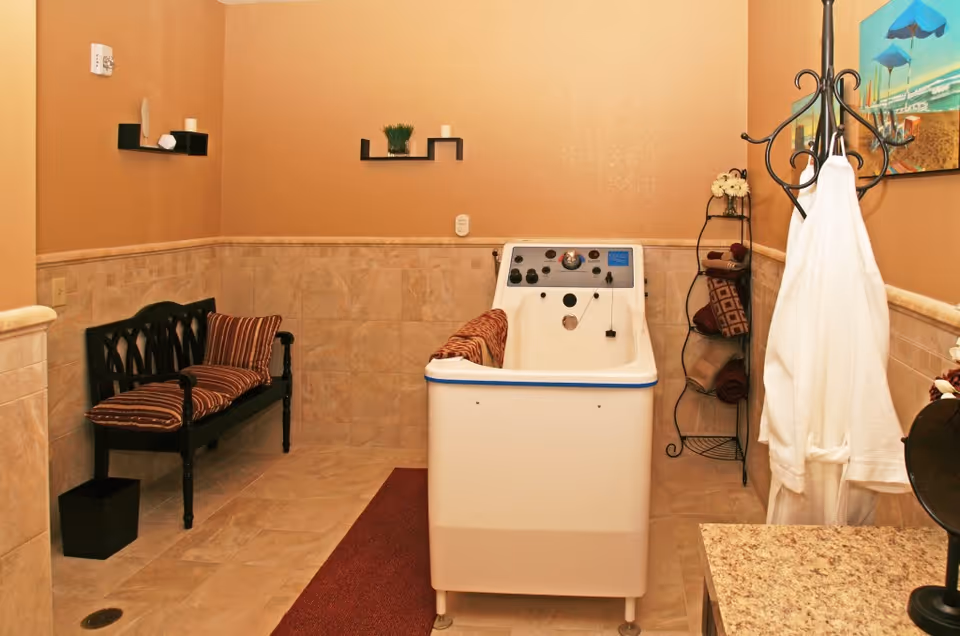 A bathing room with a white walk-in bathtub in the center, a black bench with striped cushions on the left, and a black metal rack holding rolled towels on the right. The walls are beige with tile halfway up, and there are small decorative shelves and a beach-themed painting on the walls. A white robe hangs on a black coat rack near the bathtub.