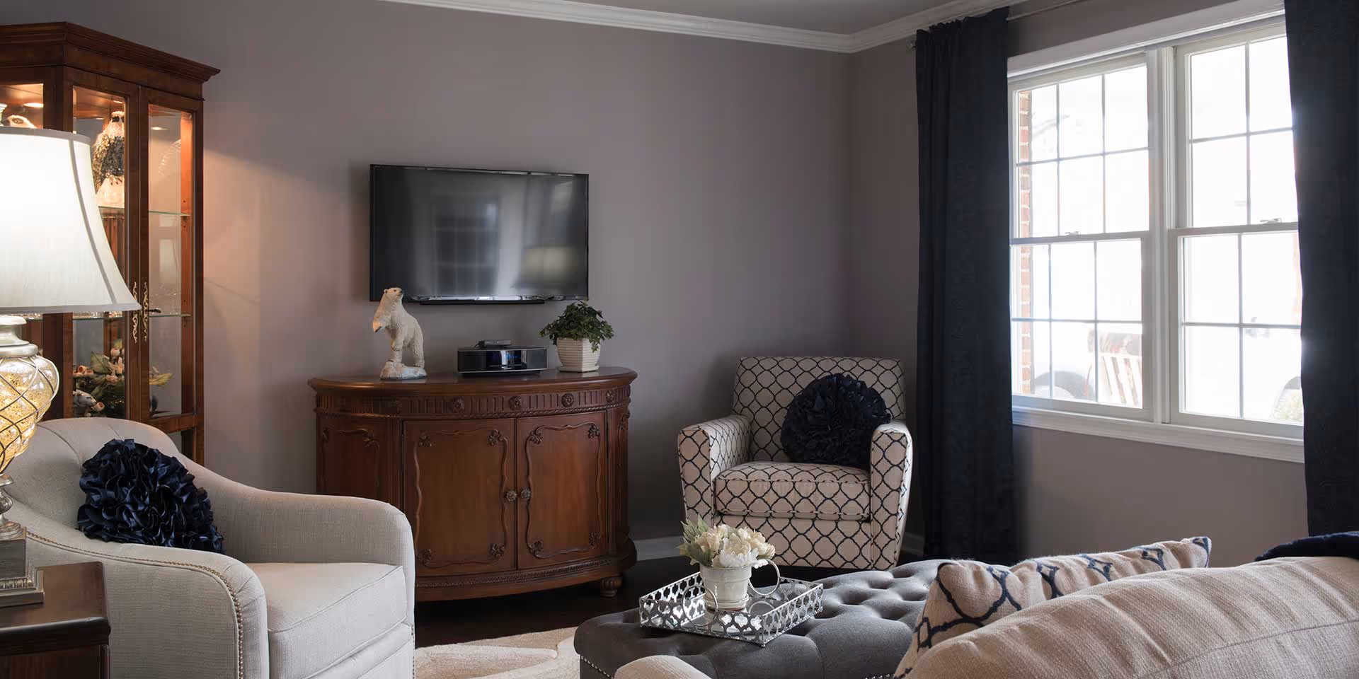 A cozy living room with a beige armchair and a patterned armchair, both with dark decorative pillows. A wooden cabinet with a small sculpture and a potted plant sits beneath a wall-mounted flat-screen TV. A tufted ottoman with a silver tray holding a small flower arrangement is in the center. Large windows with dark curtains allow natural light into the room.