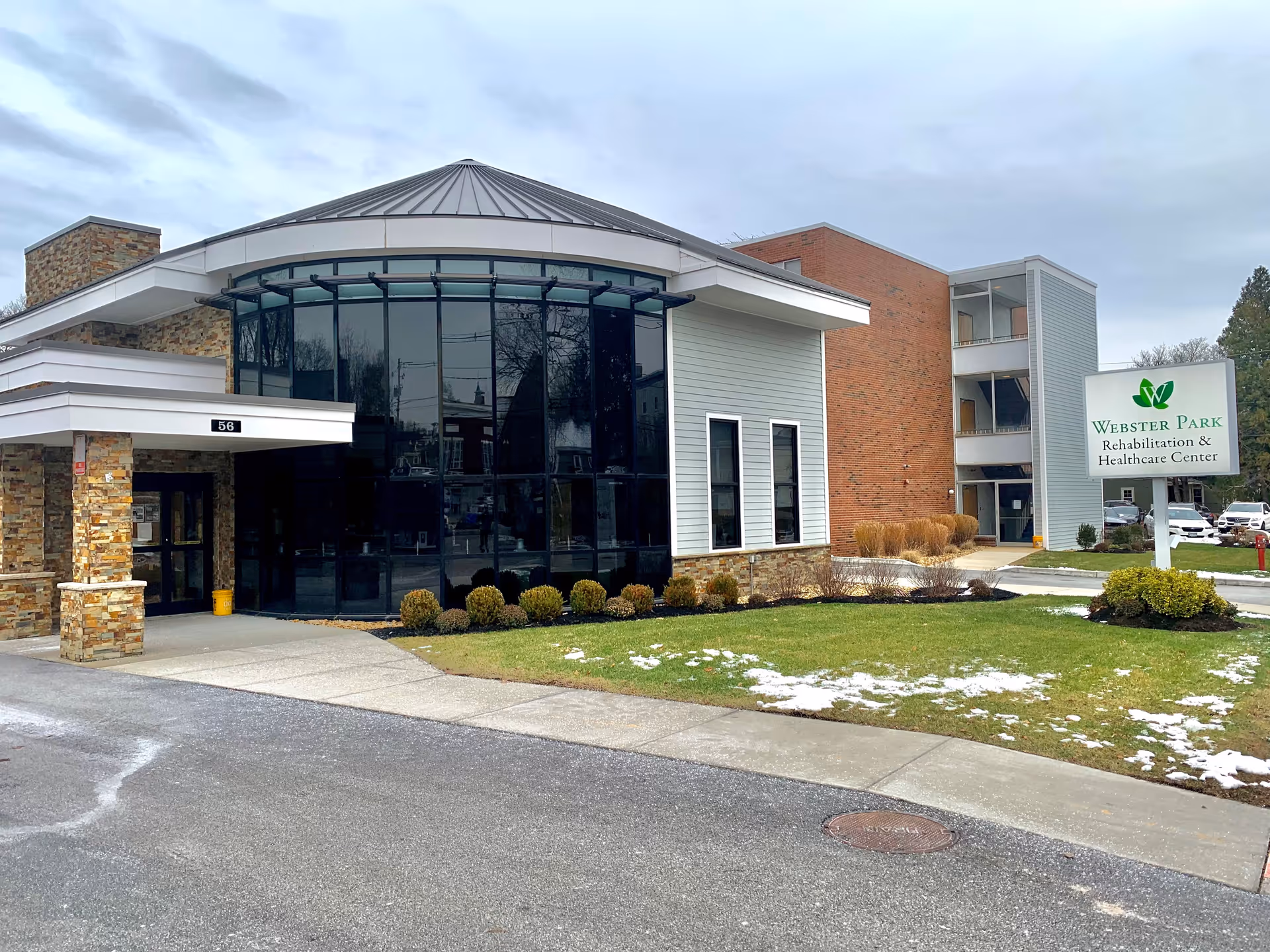 Front entrance of Webster Park Rehabilitation & Healthcare Center with a curved glass facade, covered driveway, and a lawn with a sign on a cloudy day.