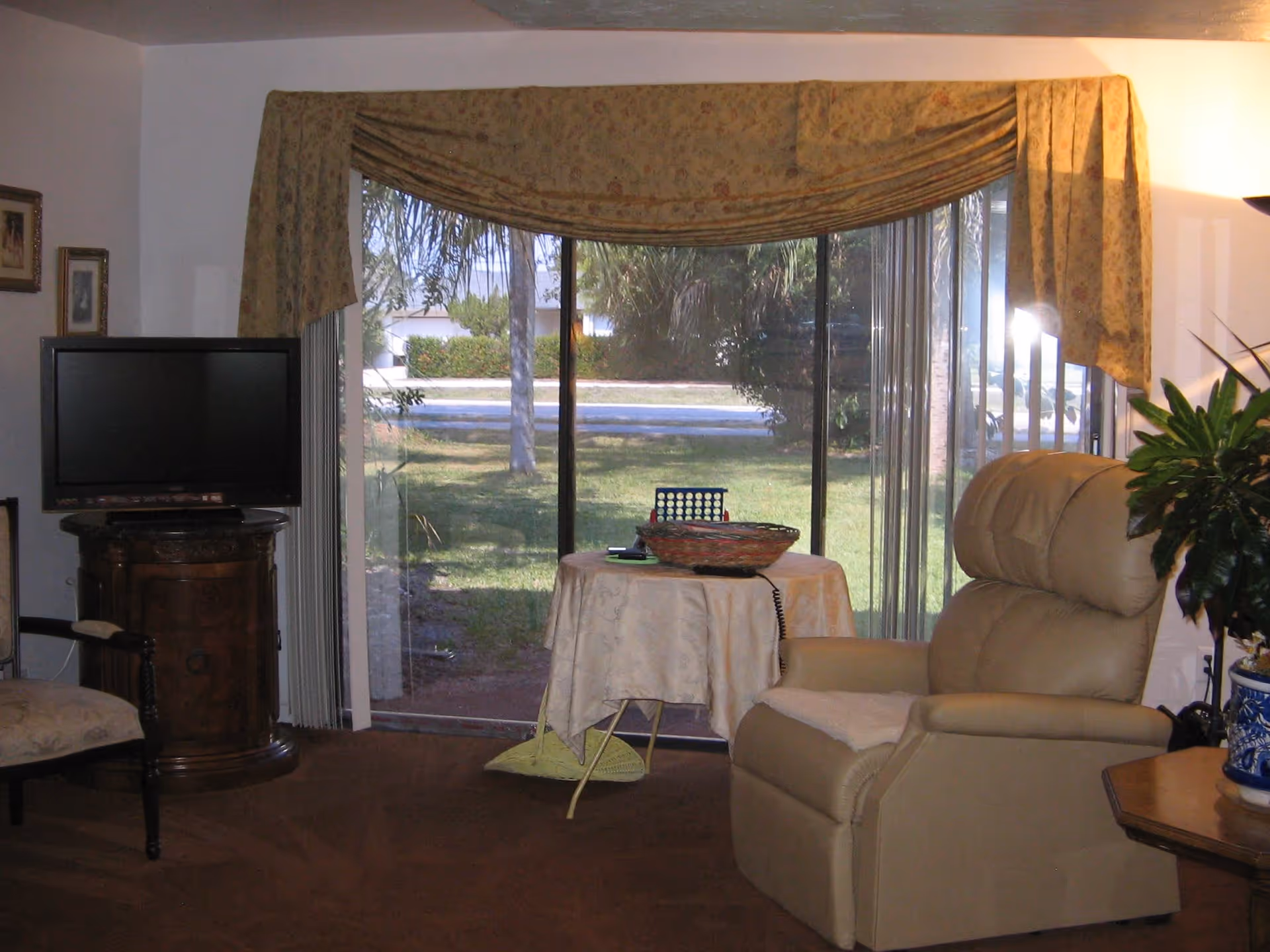 A cozy living room with a large beige recliner chair, a small round table covered with a tablecloth holding a basket and a game, a flat-screen TV on a wooden stand, a plant in a blue and white pot on a side table, and a large sliding glass door with floral curtains showing a green outdoor area.