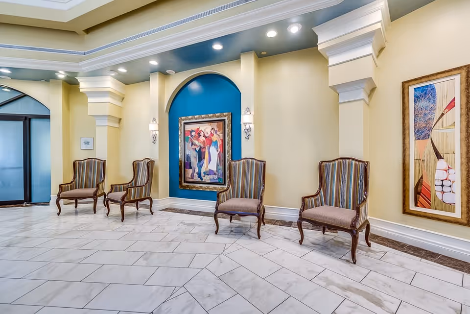 A bright, elegant waiting area with four striped upholstered armchairs arranged along a cream-colored wall. The wall features two framed colorful paintings, one set in a blue recessed arch with wall sconces on either side. The floor is covered with white marble tiles, and the ceiling has recessed lighting and decorative molding.
