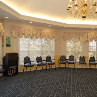 A spacious room with a patterned carpet and a row of black chairs lined up against the walls under large windows with beige valances. A small TV sits on a stand in the corner, and a chandelier hangs from the ceiling.