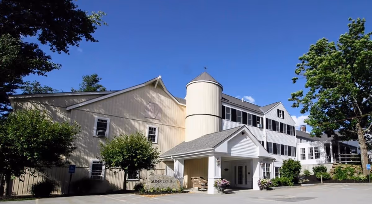Exterior view of a senior living facility building named Summerhill, featuring a white and beige structure with multiple windows, a small covered entrance with seating, surrounded by trees and clear blue sky.