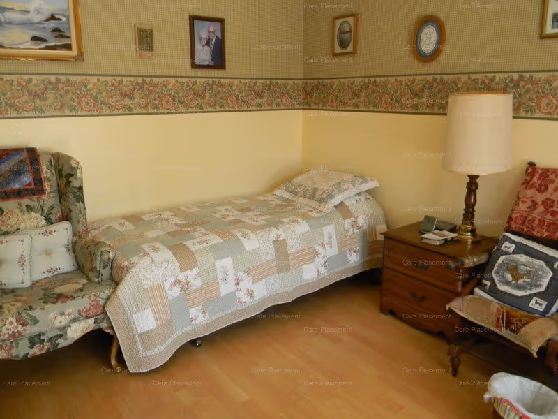 Small furnished bedroom with a single bed covered by a patchwork quilt, two armchairs, a nightstand and lamp against a floral wallpapered wall.