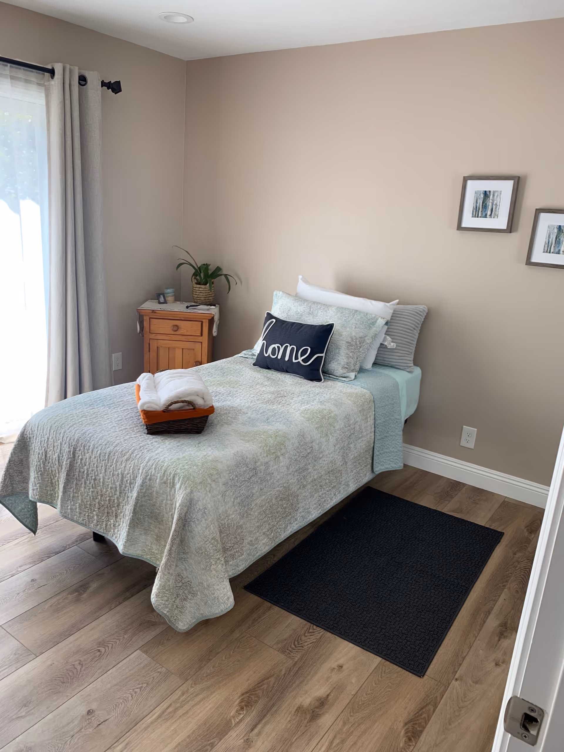 A neatly made single bed with a light-colored quilt and multiple pillows, including one with the word 'home' on it. A small wooden nightstand with a plant and some items is beside the bed. The room has light beige walls, two framed pictures on the wall, wood flooring, a black rug beside the bed, and a window with light curtains letting in natural light.