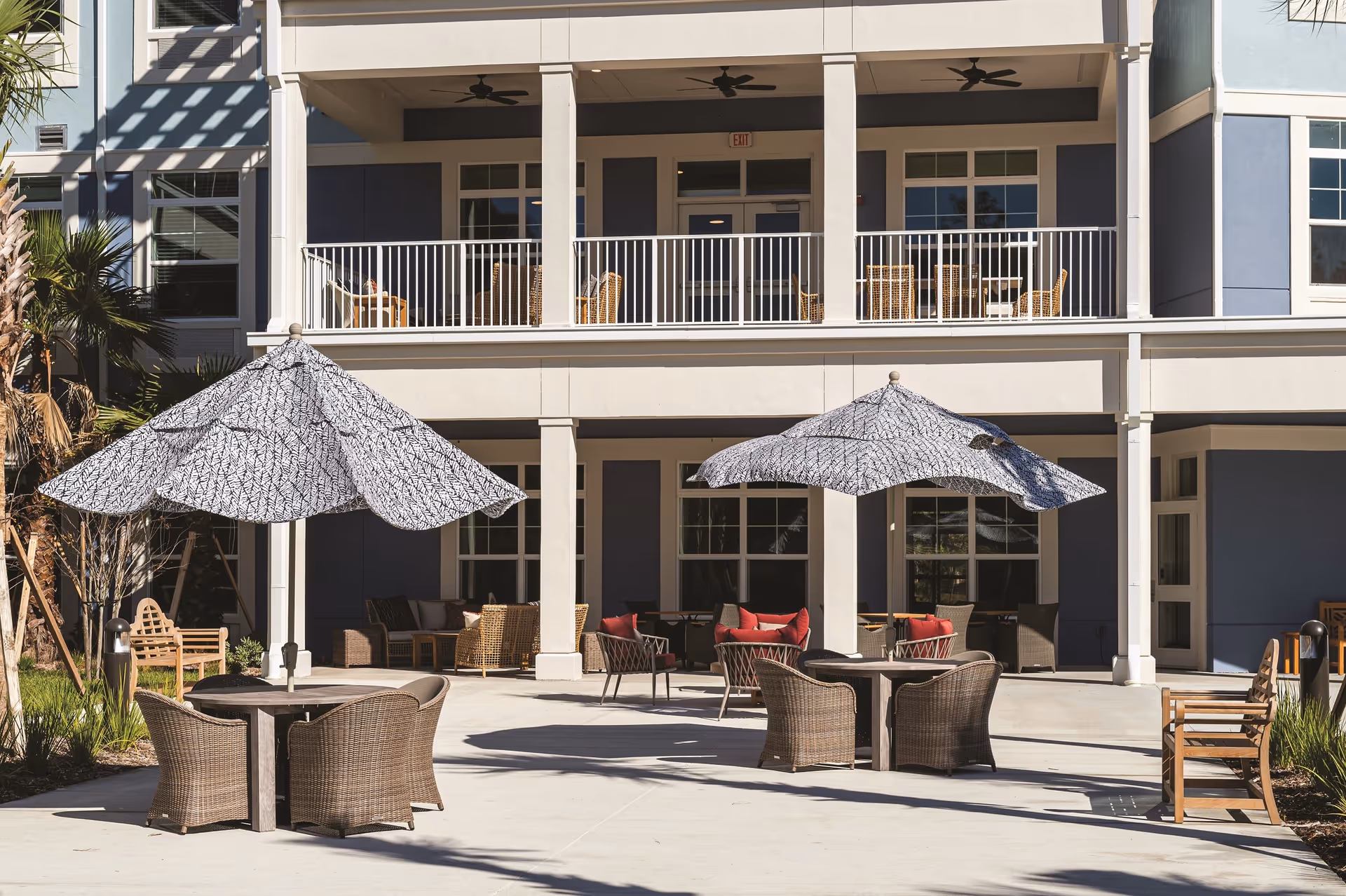 Outdoor patio area at Sancerre at Palm Coast with tables and wicker chairs under large patterned umbrellas. The patio is in front of a building with a balcony featuring additional seating and ceiling fans.