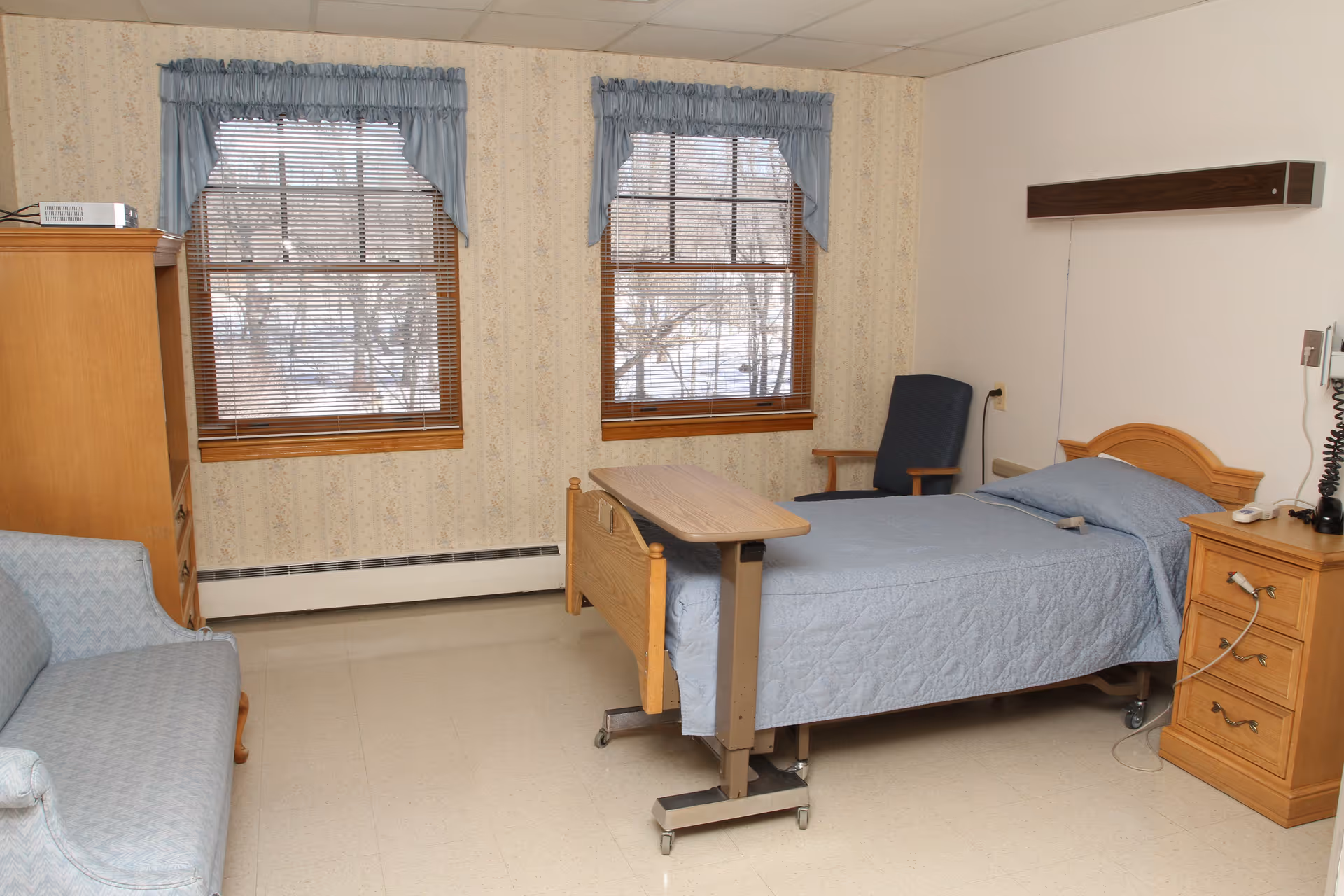 A simple, clean room with a single hospital bed covered in a blue quilt, a wooden bedside table with medical equipment, a blue armchair, a light blue couch, two windows with blue valances, and beige wallpaper with a subtle floral pattern.