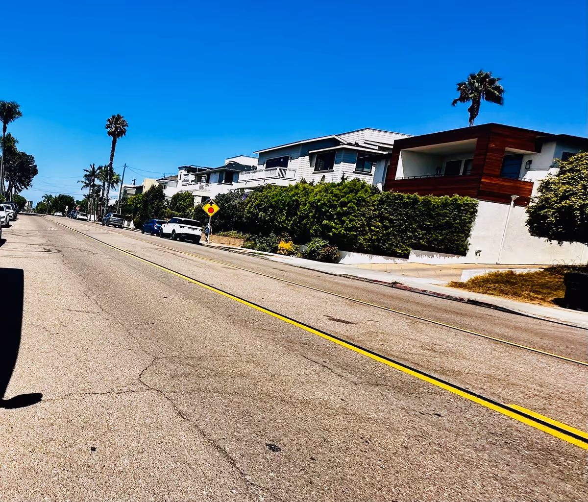 Sunny coastal street lined with modern houses, palm trees, parked cars, and a double yellow center line.