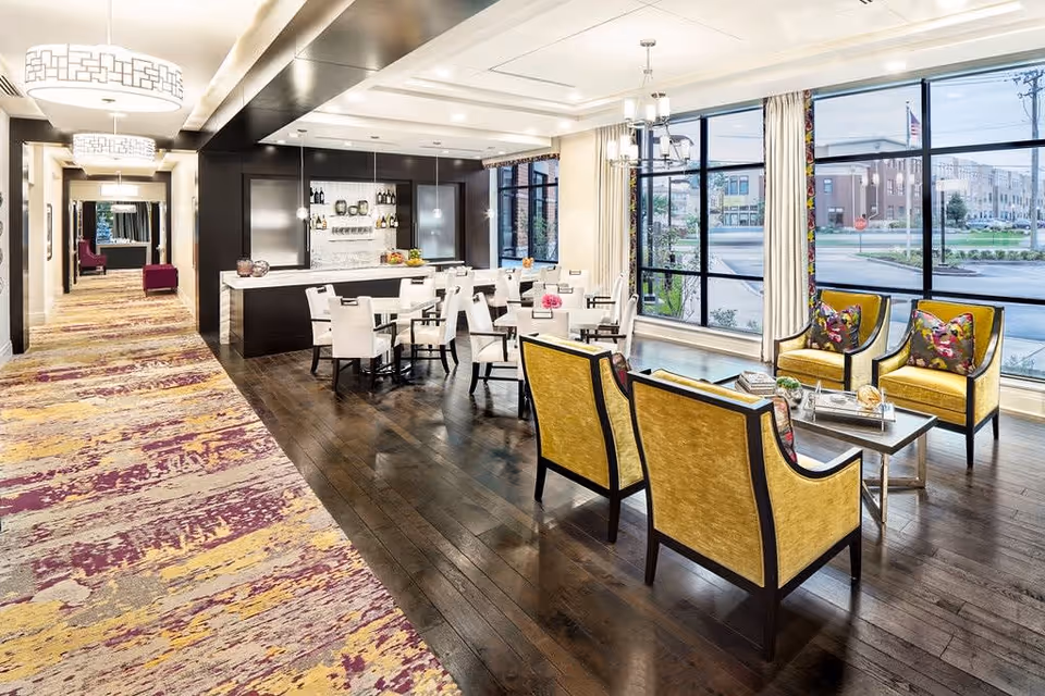 A bright and modern common area in a senior living facility featuring a seating area with yellow armchairs and a coffee table, a dining area with white chairs and tables, large windows letting in natural light, and a hallway with patterned carpet and decorative ceiling lights.