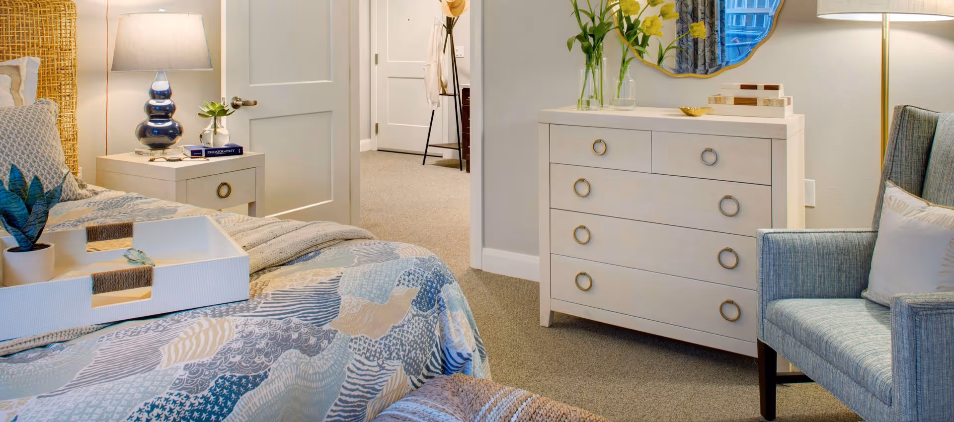 A cozy bedroom featuring a bed with a patterned quilt and a white tray holding a small plant and glasses. Next to the bed is a nightstand with a blue lamp and a book. Across from the bed is a white dresser with round handles, decorated with vases of yellow flowers and a mirror above it. A comfortable blue armchair with a white pillow sits nearby. The room has beige carpeting and light-colored walls, with an open door leading to another room.