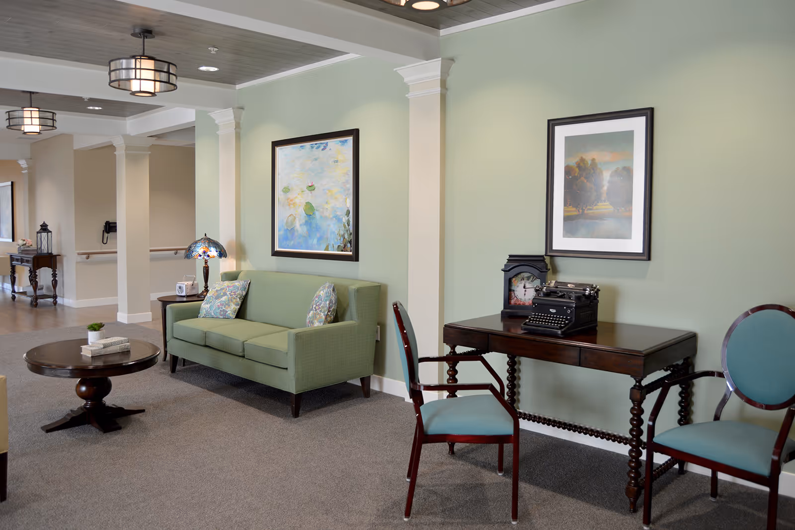A cozy interior common area with a green sofa, two floral pillows, a round wooden coffee table with books and a small plant, and a wooden side table with a decorative lamp. There is a wooden desk with an antique typewriter and a clock, flanked by two blue upholstered chairs. The walls are painted light green and decorated with framed artwork. The ceiling has modern light fixtures and white columns separate the space.