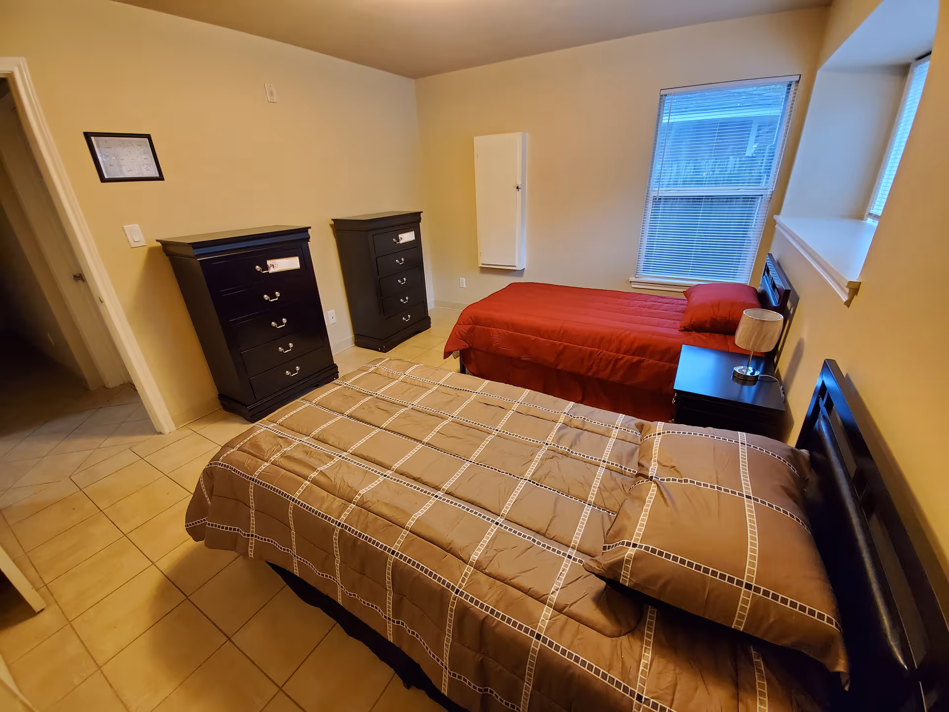 A bedroom with two single beds, one with a brown checkered comforter and the other with a red comforter. There are two black dressers against the wall, a nightstand with a lamp between the beds, and a window with blinds letting in natural light.