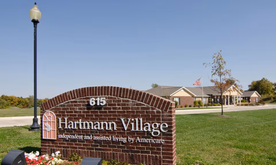Brick entrance sign reading "615 Hartmann Village" on a lawn with the senior living building and an American flag visible in the background.