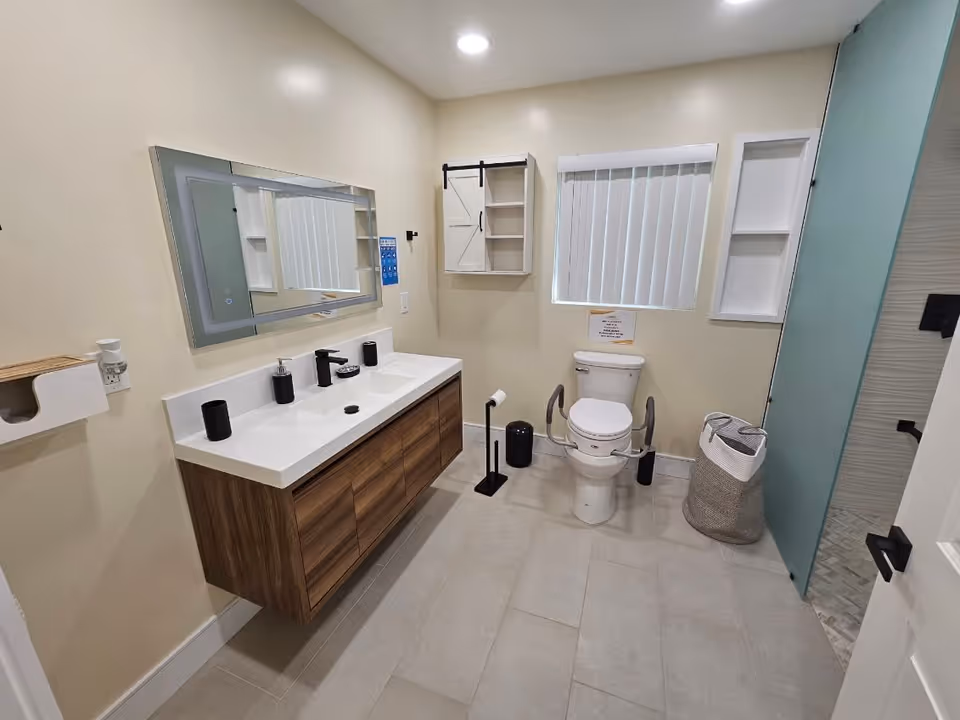 A modern bathroom with a double sink vanity featuring black fixtures and accessories. There is a large mirror above the sinks, a toilet with safety rails, a window with vertical blinds, a wall-mounted cabinet, and a laundry basket. The shower area has a frosted glass door with black hardware.