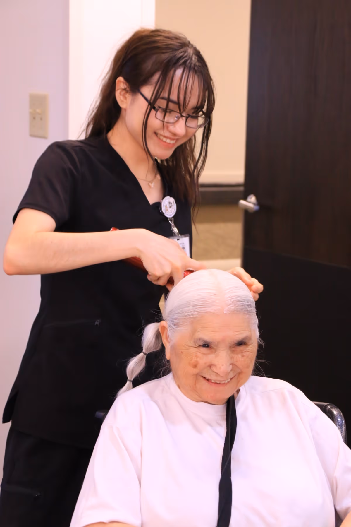 A young caregiver with glasses and a black uniform is smiling while brushing the hair of an elderly woman with white hair tied in a braid, who is seated and smiling.