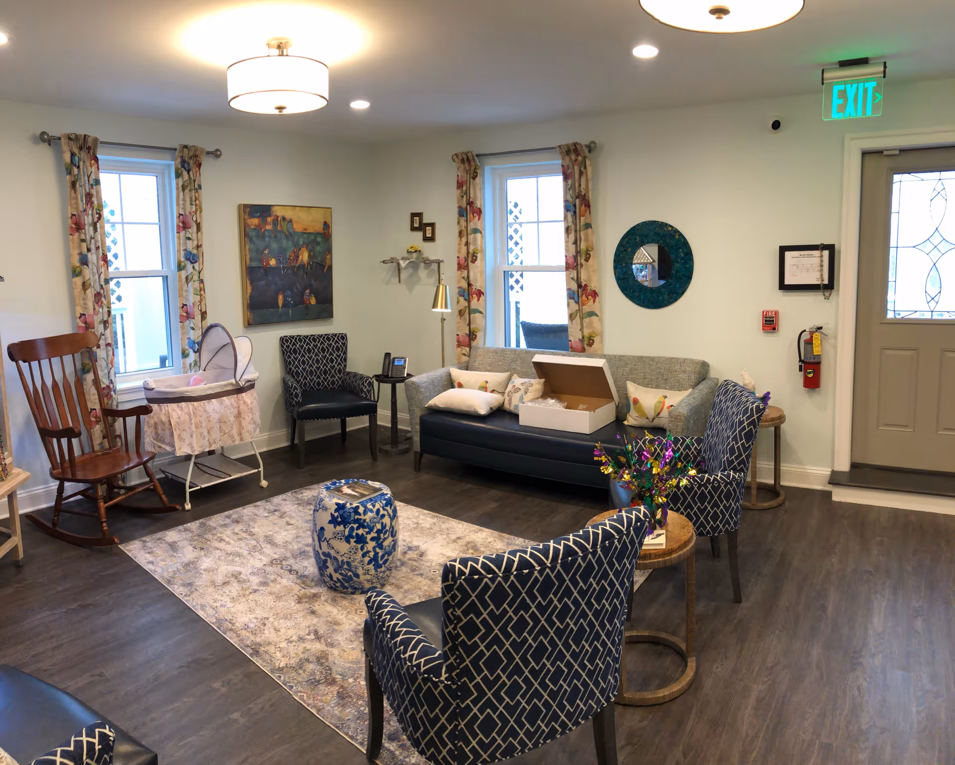 A cozy living room with a gray sofa adorned with pillows, two patterned armchairs, a wooden rocking chair, and a baby bassinet. The room has two windows with floral curtains, a round blue decorative mirror on the wall, a painting, and a small table with a colorful floral arrangement. The floor is dark wood with a light area rug, and there is a door with a glass panel and an illuminated exit sign above it.