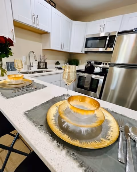 Modern kitchen with white cabinets, stainless steel appliances including a refrigerator, stove, and microwave. A white countertop island is set with two place settings featuring gold-rimmed plates, bowls, and glasses on gray placemats. A vase with red flowers is visible on the left side near the sink.