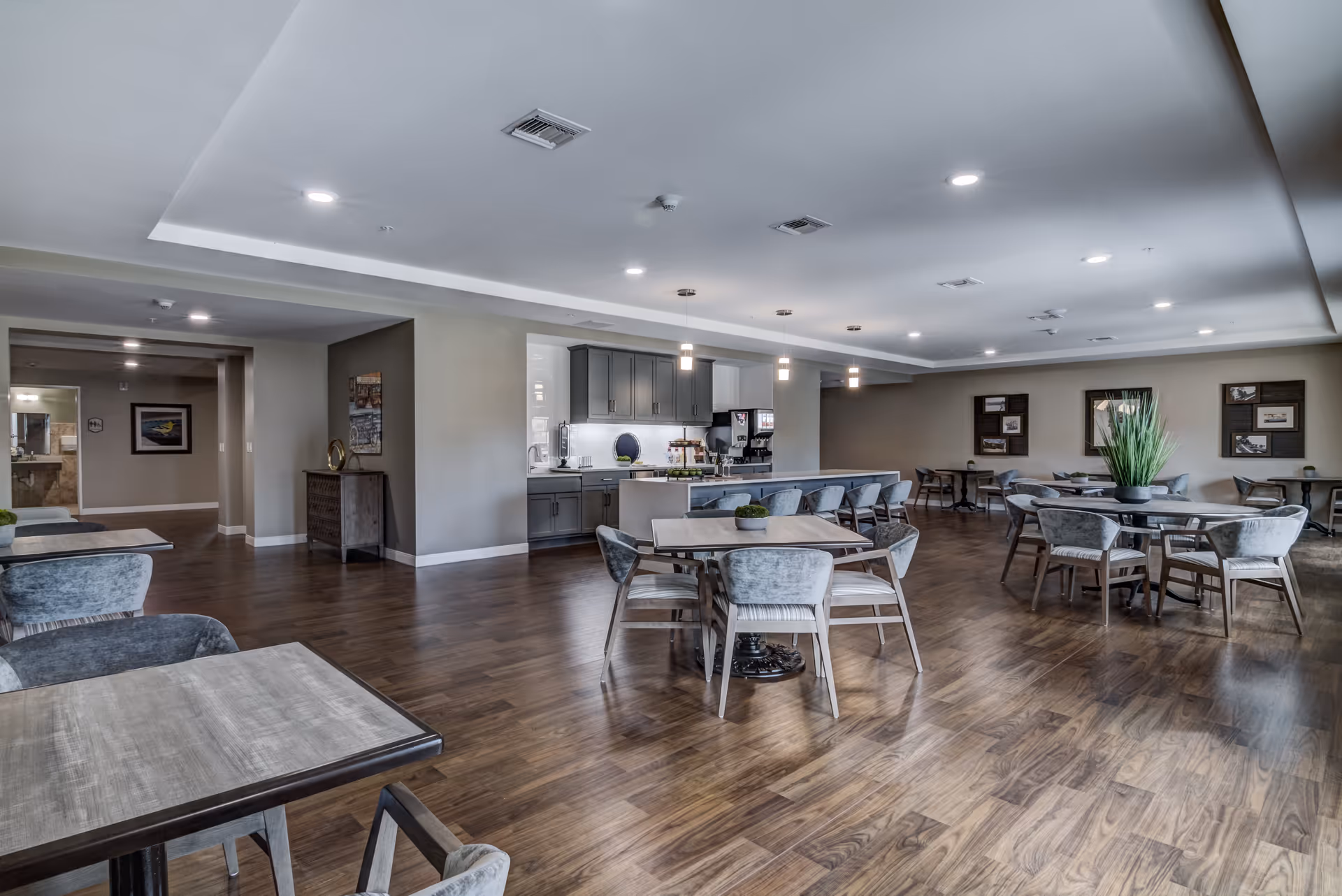 A spacious and modern dining area in an assisted living facility with multiple tables and chairs arranged on a wooden floor. The room features a small kitchen area with gray cabinets, a countertop with bar stools, and pendant lights hanging from the ceiling. The walls are decorated with framed pictures and a large plant is placed on one of the tables.