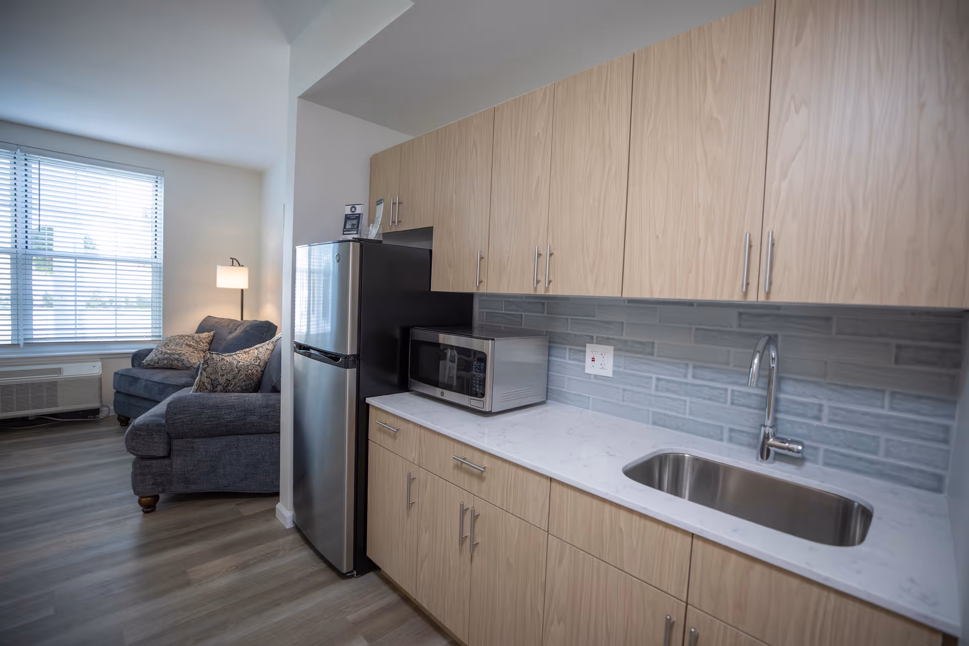 A modern kitchenette with light wood cabinets, a stainless steel refrigerator, a microwave on the counter, and a small stainless steel sink. In the background, there is a cozy living area with a dark gray armchair, a floor lamp, and a window with blinds letting in natural light.