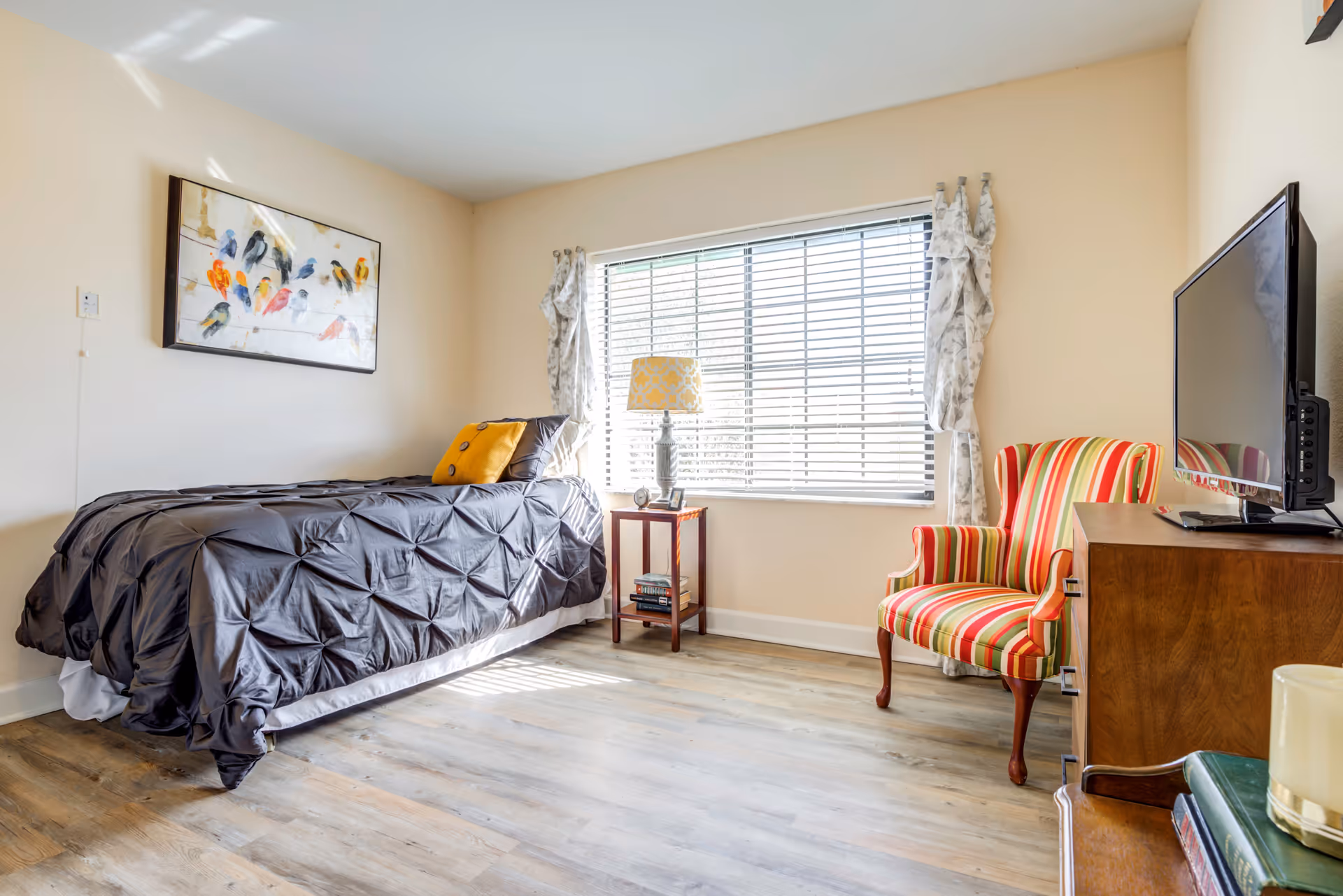 A bright bedroom with a single bed covered in a dark gray comforter and a yellow pillow. Next to the bed is a small wooden nightstand with a lamp and some books. A large window with white blinds and patterned curtains lets in natural light. Across from the bed is a wooden dresser with a flat-screen TV on top and a striped armchair with red, yellow, and green colors beside it. The floor is light wood, and a colorful bird painting hangs on the wall above the bed.