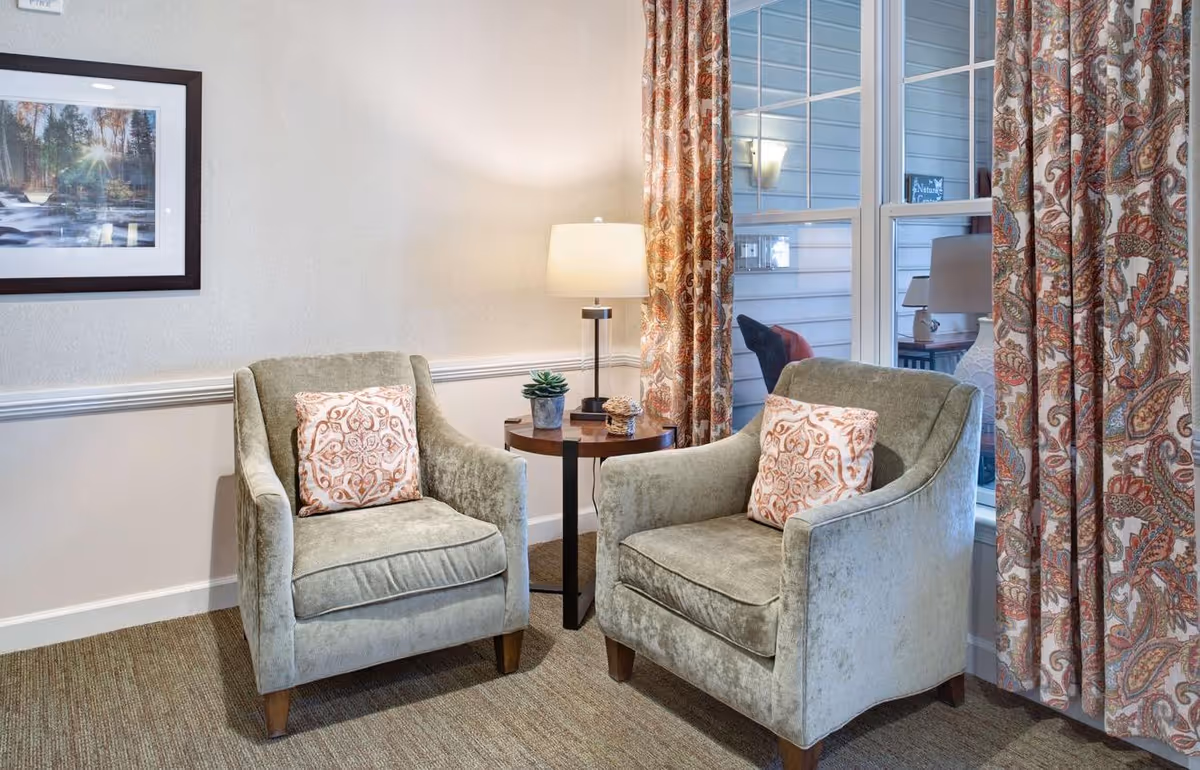 Two upholstered armchairs with patterned pillows flank a small round side table and lamp in a cozy sitting area with patterned curtains and a framed picture on the wall.