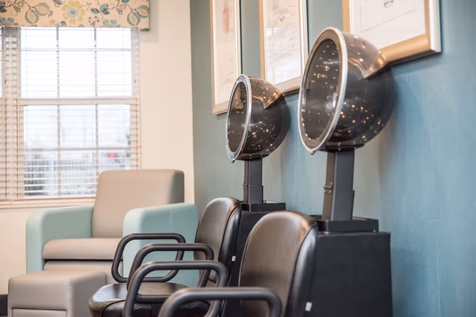 Interior view of a salon area with two black hair drying chairs and hair dryers mounted on stands against a teal wall. In the background, there is a light gray armchair with light blue armrests and a window with blinds and floral valance.