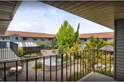 View from a covered balcony overlooking a landscaped courtyard with a pond, trees, shrubs, and a gazebo, surrounded by a two-story building with a red roof under a partly cloudy sky.