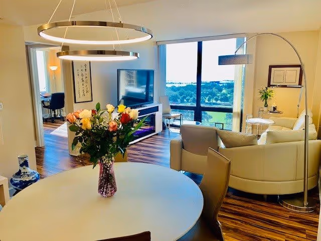 A bright and modern living area with a round dining table in the foreground holding a vase of colorful roses. The room features wood flooring, a large window with a scenic view, a curved white sofa, a floor lamp, a flat-screen TV on a stand, and a small desk with a chair in an adjacent room.