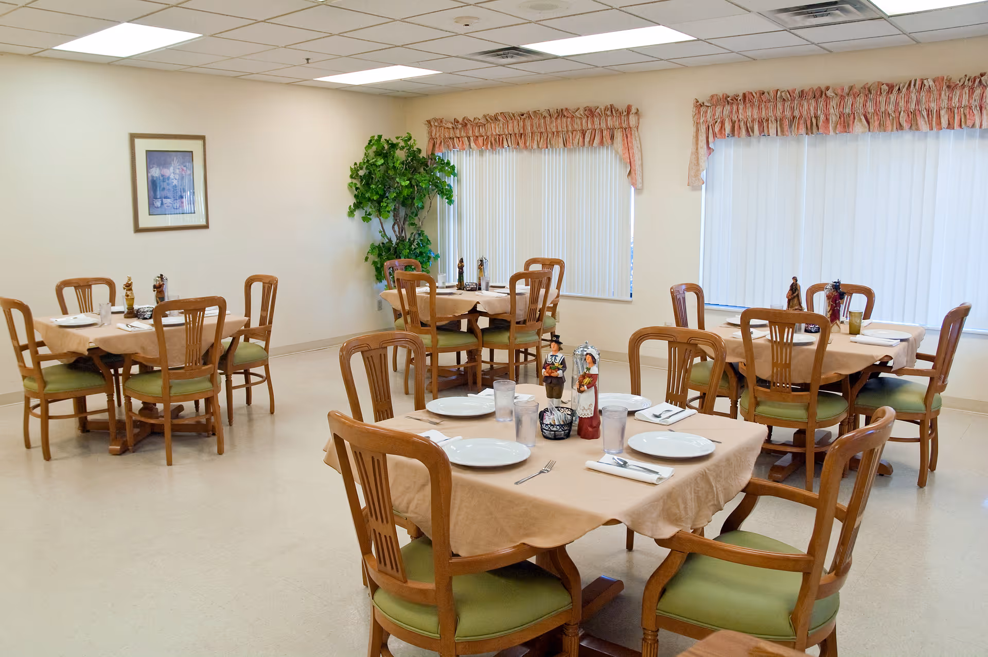 Bright institutional dining room with multiple tables set for a meal, wooden chairs, and large windows with vertical blinds.