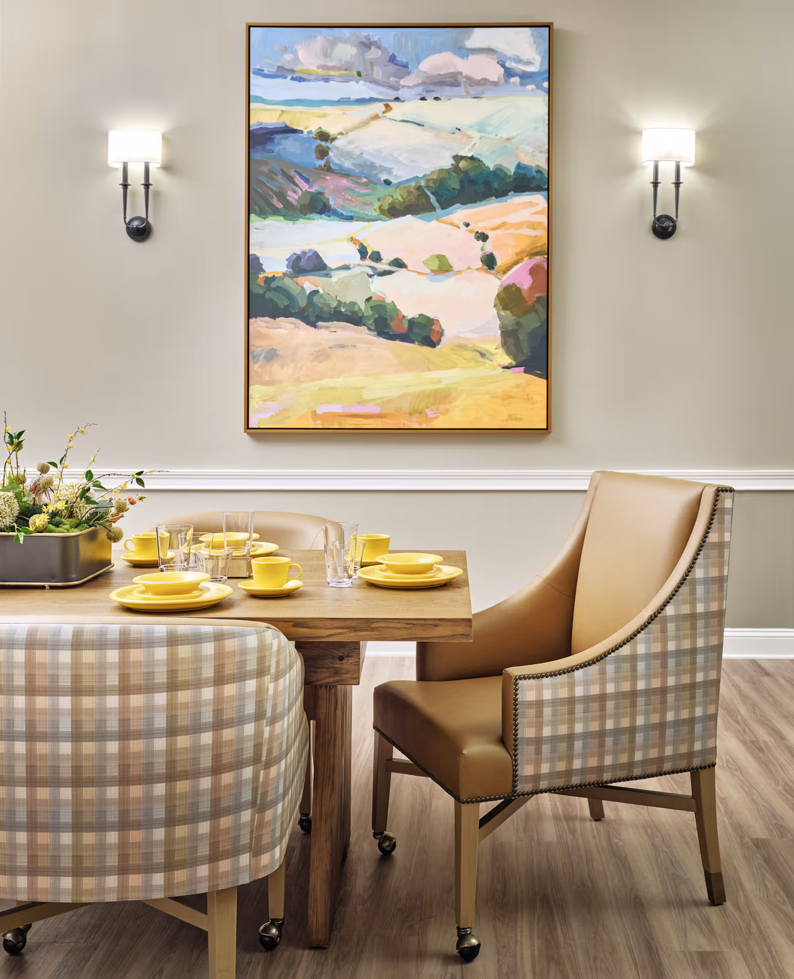 A dining area with a wooden table set with yellow plates, cups, and clear glasses. The chairs around the table have a plaid pattern with beige and brown tones, and one chair has leather upholstery. On the wall above the table is a colorful landscape painting, flanked by two wall-mounted light fixtures. The floor is wood, and there is a decorative plant centerpiece on the table.