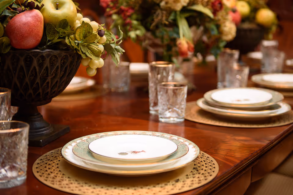 Close-up view of a dining table set with decorative plates, clear drinking glasses, and a centerpiece featuring artificial fruit and greenery in a dark ornamental vase.