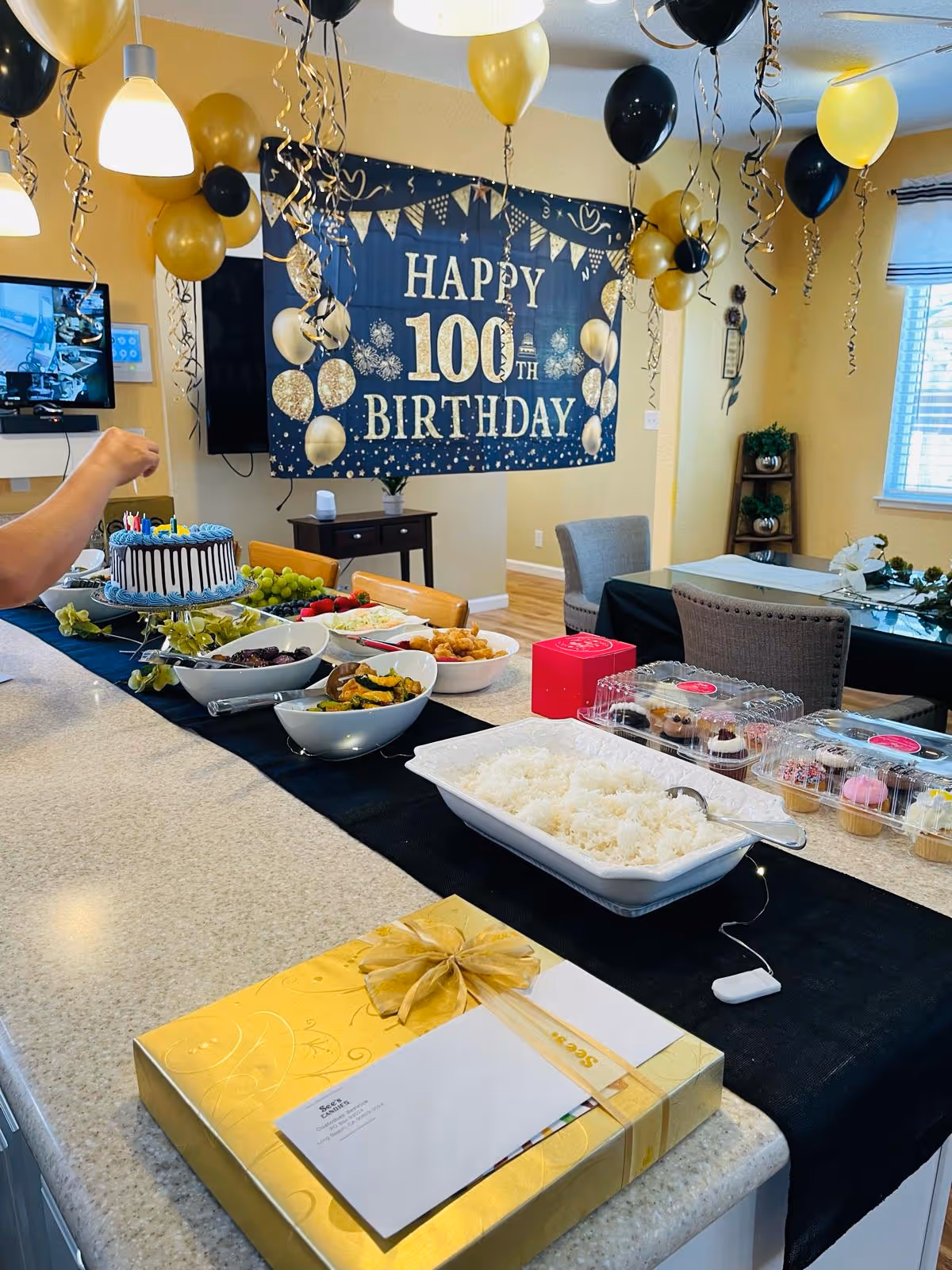 A decorated dining area with a banner reading 'Happy 100th Birthday' in gold and black colors. The room has black and gold balloons hanging from the ceiling. A table is set with various dishes including rice, cupcakes, and other food items. A golden gift box with a ribbon and an envelope is placed on the counter in the foreground. A person’s arm is visible reaching towards a birthday cake with candles.