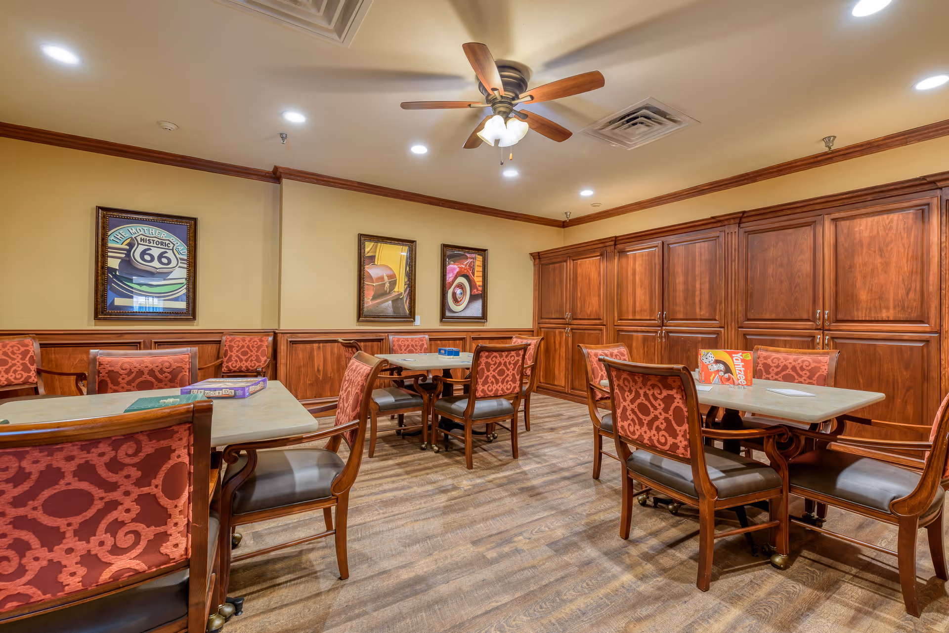 A cozy room with several tables and chairs featuring red patterned upholstery. The room has wooden cabinets along one wall, framed vintage car pictures on the walls, a ceiling fan with lights, and recessed ceiling lights. The floor is wood, and the room appears well-lit and inviting.