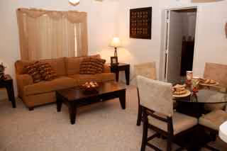 A living room and dining area in Heron Pond Apartments featuring a beige sofa with patterned pillows, a dark wooden coffee table with a decorative bowl, a side table with a lamp, and a round glass dining table with four cushioned chairs set with plates and cups. The walls are light-colored with a window covered by beige curtains and a framed decorative piece.