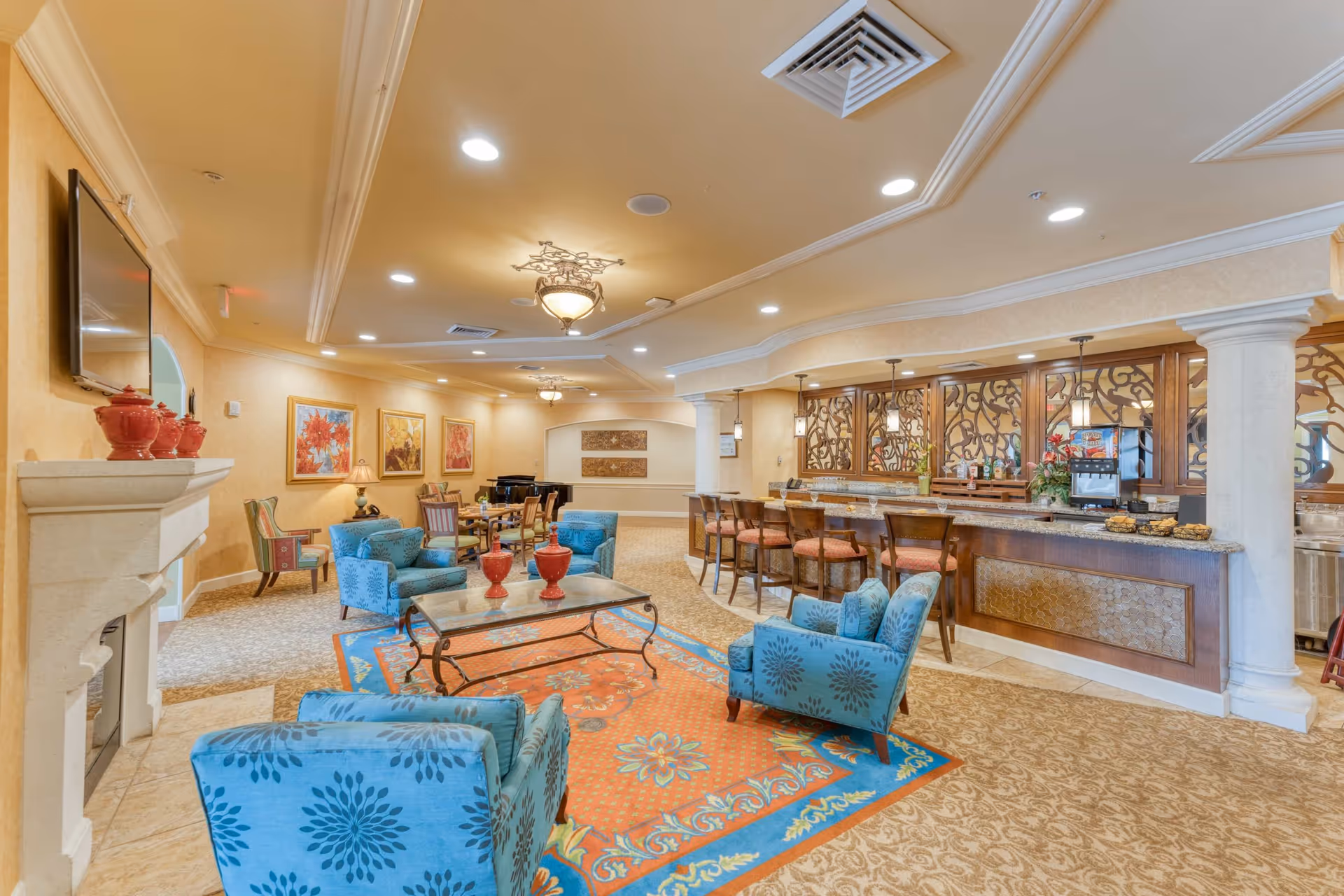 A spacious and elegantly decorated common area in a senior living facility featuring blue patterned armchairs around a glass coffee table on a colorful rug, a fireplace with red decorative vases, a bar counter with high chairs, and framed floral artwork on the walls under warm ceiling lights.