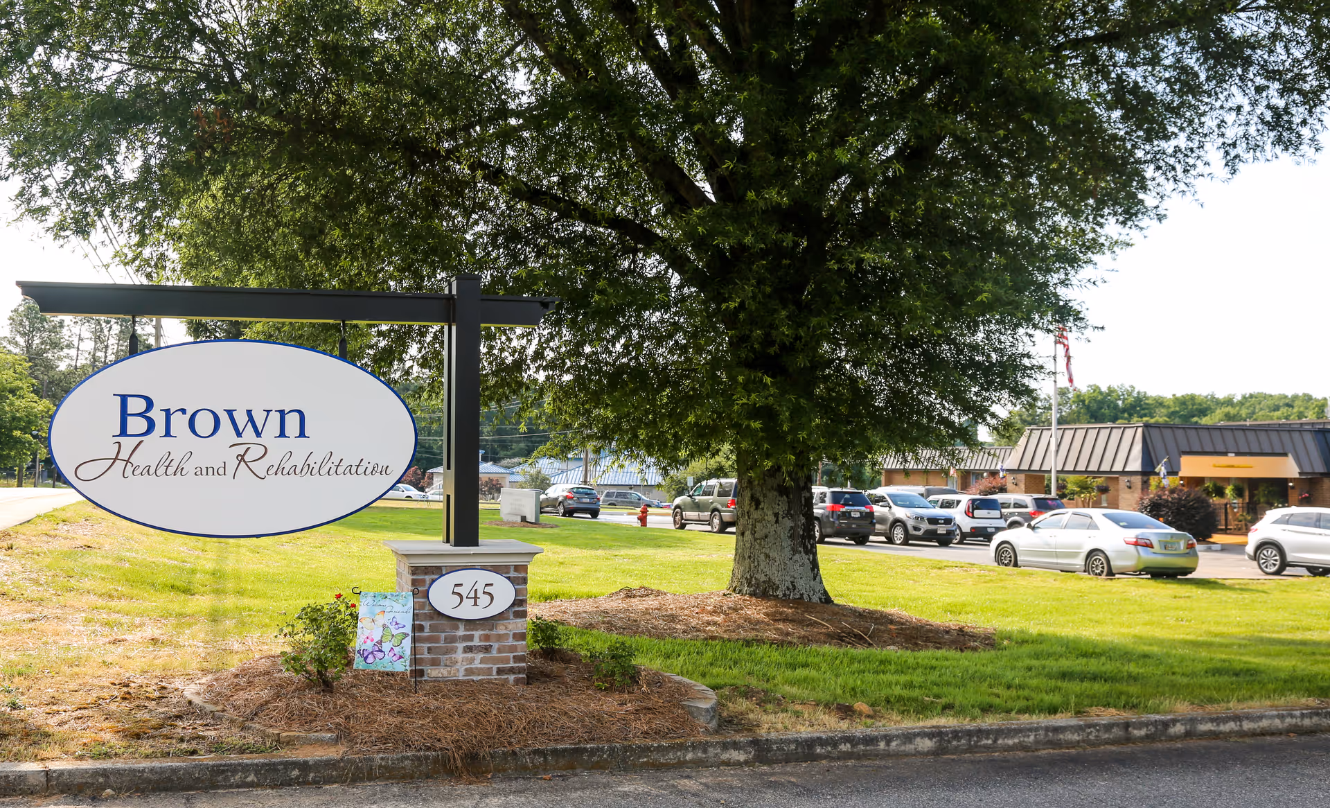 Outdoor view of Brown Health and Rehabilitation facility sign on a brick post with the number 545, a large tree, a grassy area, and a parking lot with several cars in front of a single-story building.