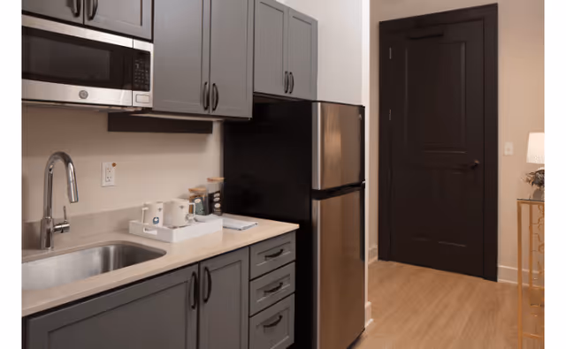 A modern kitchenette featuring gray cabinets, a stainless steel microwave mounted above the counter, a stainless steel refrigerator, a sink with a curved faucet, and a countertop with a tray holding coffee mugs and supplies. A dark-colored door and a small table with a lamp are visible in the background.