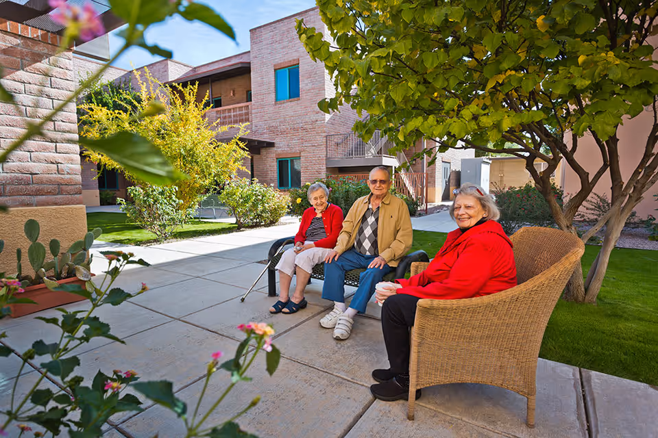 Three elderly people sitting outdoors in a courtyard area of a senior living facility. Two women and one man are seated on chairs and a bench, surrounded by greenery, trees, and brick buildings under a clear blue sky.