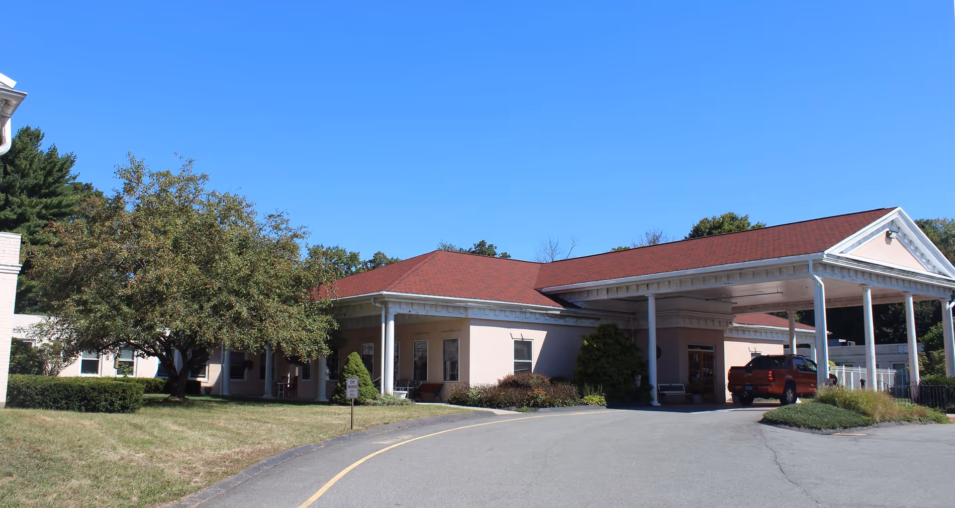 Exterior view of Autumn Lake Healthcare at Bucks Hill facility showing a single-story building with a red roof, a covered entrance with white pillars, a driveway, and surrounding greenery including trees and bushes under a clear blue sky.