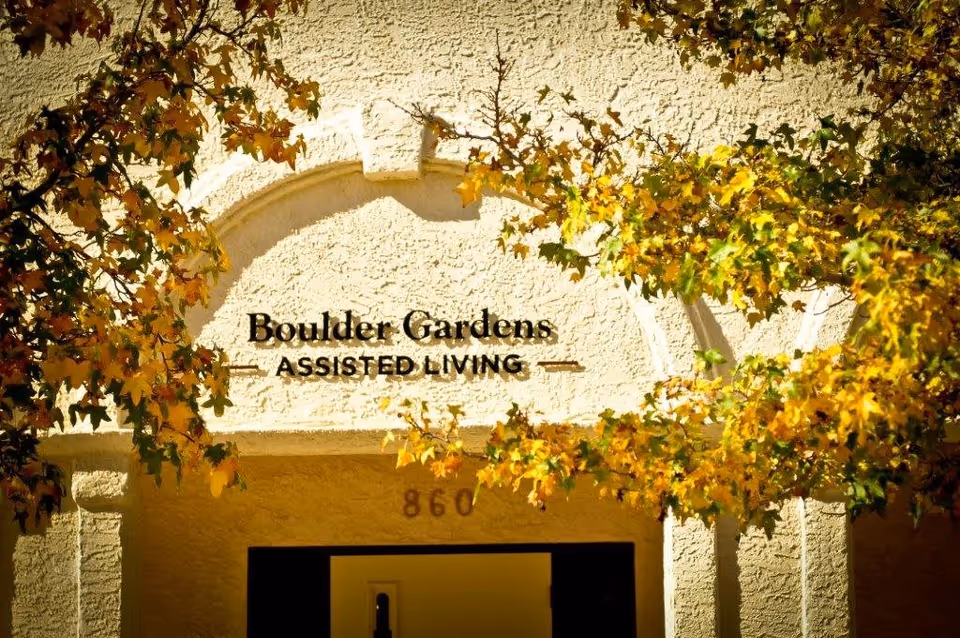 Entrance of Boulder Gardens Assisted Living facility with textured beige walls and an archway above the door. The building is partially framed by tree branches with yellow and green leaves.