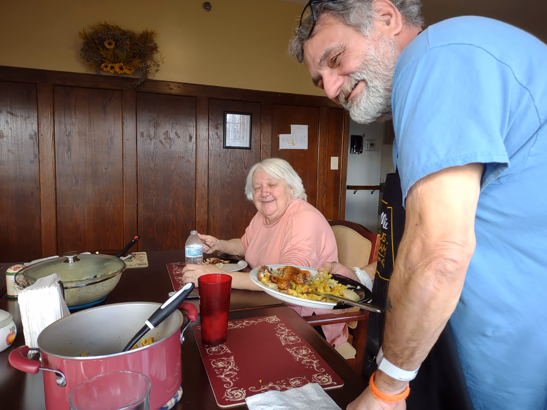 An elderly woman sitting at a dining table with a plate of food and a bottle of water, smiling as a man in a blue shirt and apron serves her more food on a plate. The room has wooden paneling on the walls and a decorative arrangement of dried flowers above.