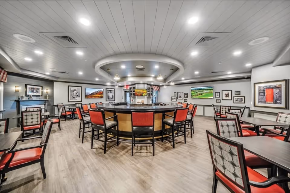 A spacious dining room with multiple tables and chairs featuring red and patterned upholstery. The room has a modern ceiling with recessed lighting and a circular central area with pendant lights. The walls are decorated with framed pictures and a flat-screen TV.