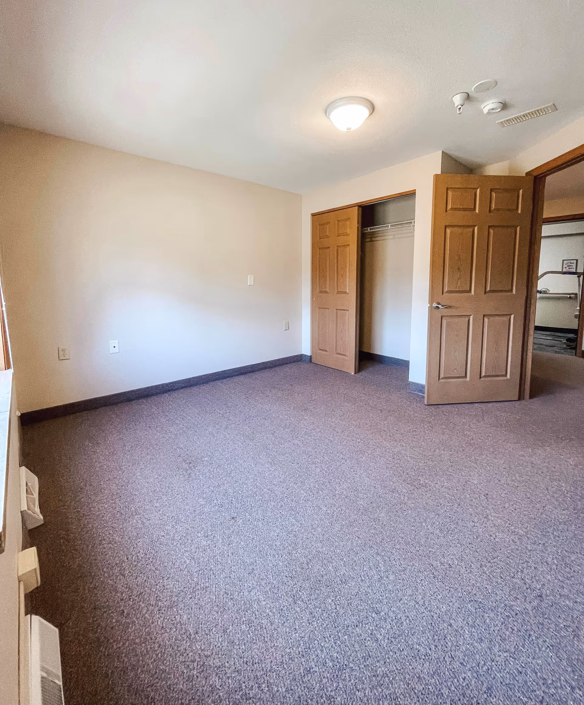 Empty room with beige walls and carpeted floor, featuring an open closet with wooden double doors and a wooden door leading to another room. Ceiling light fixture is visible.