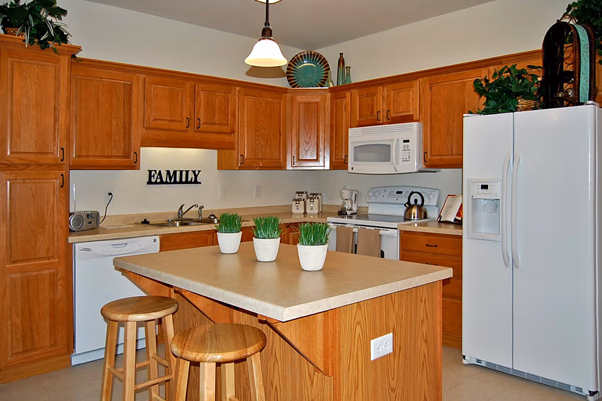 Bright kitchen with wooden cabinets, a central island topped with three potted plants and two stools, and white appliances.