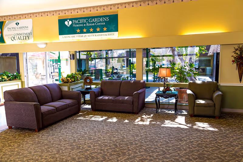 A bright and spacious seating area inside Pacific Gardens Nursing and Rehabilitation Center featuring two purple sofas and one green armchair arranged around a small glass-top table with a lamp. Large windows behind the seating area provide a view of an outdoor patio with greenery and seating. Above the windows, there are two signs displaying the facility's name and quality rating.